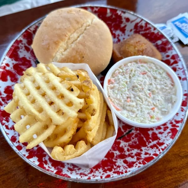 Cindy Lou's BBQ meal with waffle fries, coleslaw, a bun, and cornbread on a red and white speckled plate.