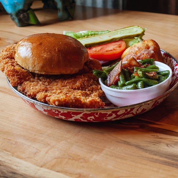Cindy Lou's BBQ fried chicken sandwich with green beans, pickles, and tomato on a rustic plate.