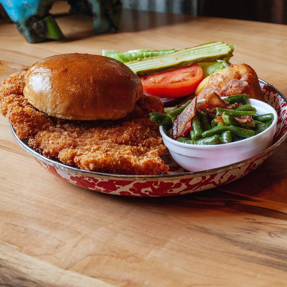 Cindy Lou's BBQ fried chicken sandwich with green beans, pickles, and tomato on a rustic plate.